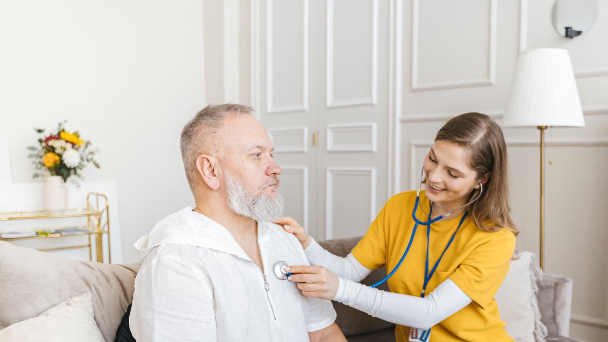 Nurse examining an older man with a stethoscope during a home visit