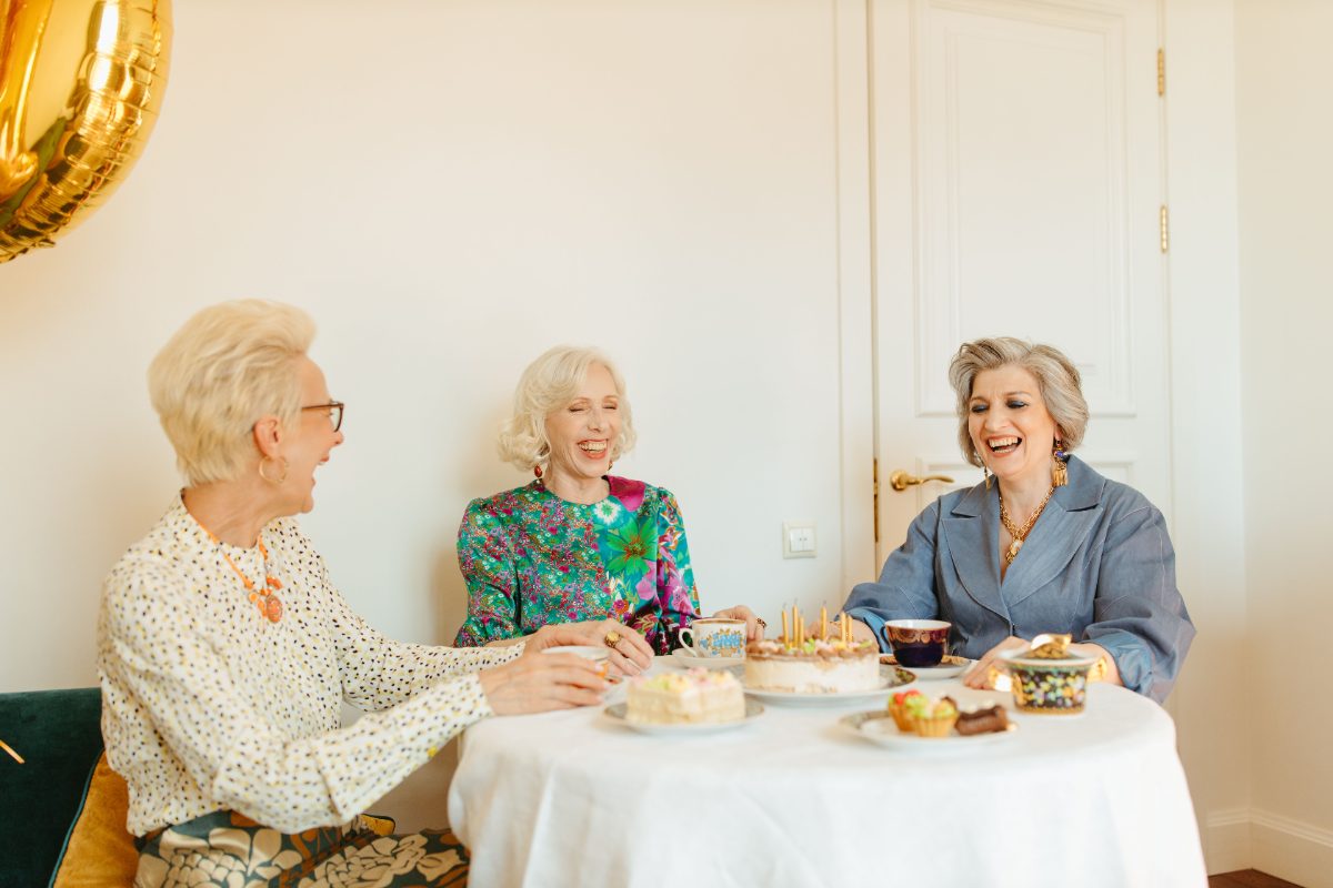 Three senior women laughing together over tea and cake at a small celebration